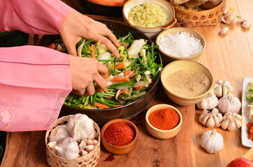 A woman wearing a pink hanbok is sitting and kneading cabbage in a large bowl to make kimchi. Nearby are various condiments such as chili powder, salt, and sugar.