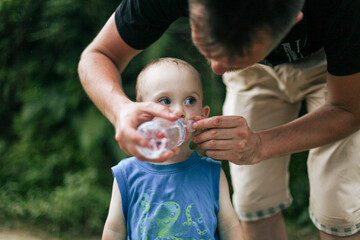 Lifestyle photo of young father gently holding bottle giving water to his tired child outdoors....