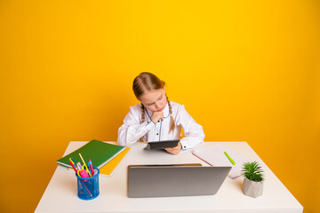 Young schoolgirl seated at desk studying with book, tablet, and laptop on yellow background