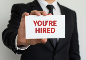 Obraz premium A person in a formal suit holds a white card with the bold red text you re hired in front of the camera