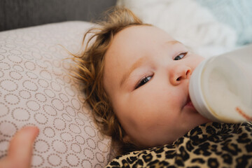 A young child with curly hair is drinking from a bottle propped up comfortably on a soft surface at...