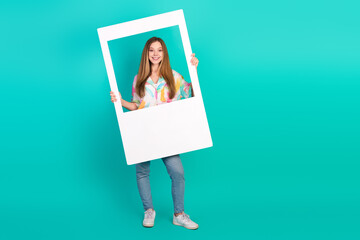 Stylish Young Woman Holding a Picture Frame on a Turquoise Background in Casual Outfit