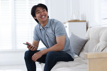 Obraz premium Smiling Asian man with smartphone on sofa at home