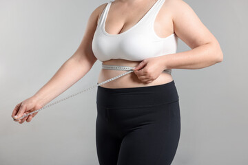 Weight loss. Plus size woman measuring waist with tape on gray background, closeup