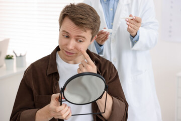 Young man putting on contact lenses near mirror in ophthalmologist office