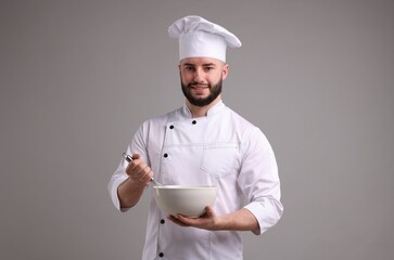 Happy confectioner in uniform holding whisk and bowl on light grey background