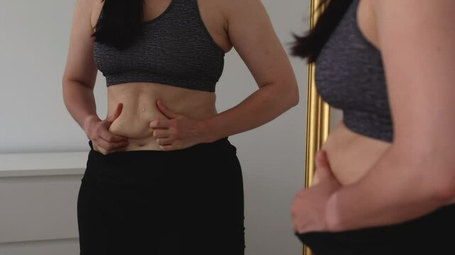 An upset woman looks at herself in the mirror, touching the fat on her stomach. She wears sportswear and stands in a bright room. The image reflects body insecurities, weight loss struggle