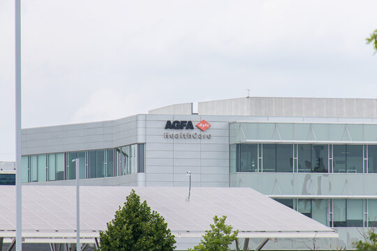 Waterloo, ON, CANADA - Aug 5 2024 : AGFA HealthCare office building with glass facade and company signage, surrounded by trees and solar panel under cloudy sky.