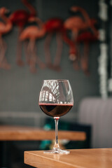 A single glass of red wine stands on a wooden table with a blurred background of pink flamingo decor and cozy indoor seating
