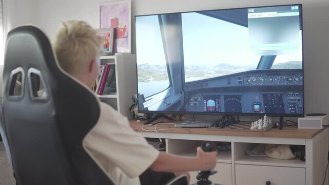 Blond boy playing a flight simulator video game at home using a gaming console and joystick. Seated in a gamer chair in front of a large TV screen displaying a realistic airplane cockpit, the child