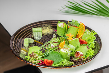 A vibrant plate of green vegetable rolls with fresh greens, cherry tomatoes, sprouts, and dried orange garnishes served on a dark ceramic dish