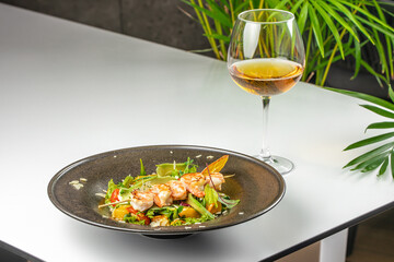 A gourmet shrimp salad with avocado, cherry tomatoes, greens, and microgreens served on a dark plate, paired with a glass of white wine