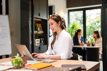 Young asian businesswoman using laptop for remote work .young woman sits at office desk , texting,...
