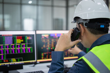 Technician in safety helmet uses computer to control systems in substation, monitoring data on multiple screens with focused attention and professional care