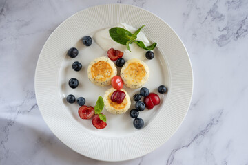 Three cheesecakes on a white plate are garnished with sour cream, blueberries, cherries and mint leaves, creating a delicious and visually appealing presentation