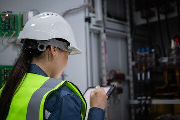 Female electrician wearing safety helmet and reflective vest performs control panel inspection using tablet while engineer monitors electrical system in industrial setting