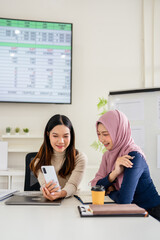 Two business woman sitting at a table in meeting room and working together. one showing her phone...