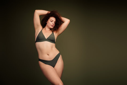 Confident young woman in olive swimwear posing against a dark green background, celebrating body positivity - Powered by Adobe