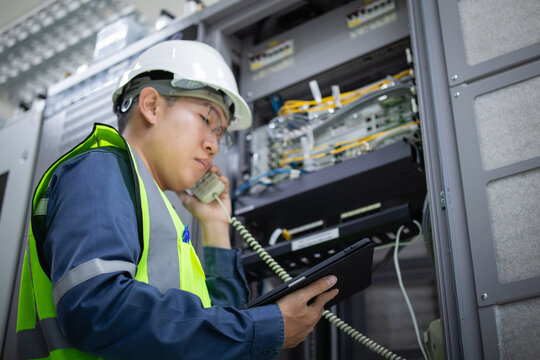Network technician wearing safety helmet and vest performs LAN patchpanel inspection while holding tablet and communicating via telephone in server room - Powered by Adobe