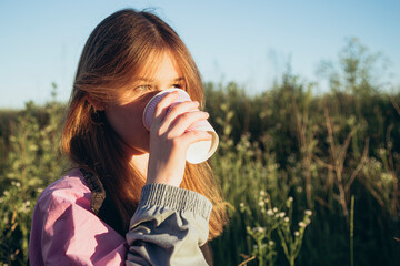 Fototapeta premium Beautiful girl drinking coffee or tea on a warm summer at sunset in a field.