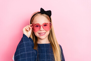 Charming young girl in a stylish outfit with heart-shaped sunglasses posing against a bright pink background with a cheerful smile