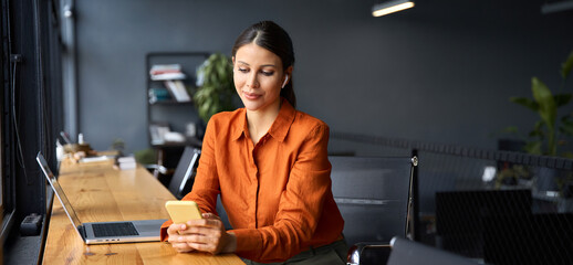Smiling Latin Hispanic female businesswoman holding smartphone in office for trading, investing, banking online. Young Indian business woman manager using cell phone mobile, laptop. Banner, copy space