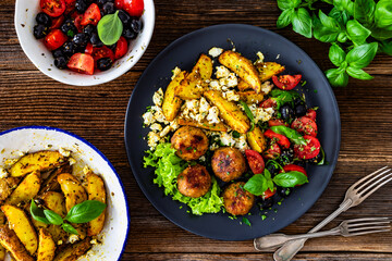 Greek style fried meatballs, fried potato wedges with feta cheese and tomato salad on wooden table	