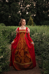 Beautiful young woman in red medieval dress is standing in the park. Fantasy princess walking in...