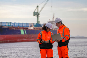Two port engineers in high-visibility orange uniforms and hard hats communicate while inspecting...