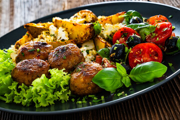 Greek style fried meatballs, fried potato wedges with feta cheese and tomato salad on wooden table	