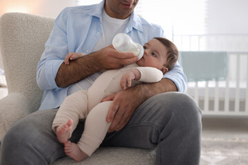 Father feeding his little baby with bottle on armchair at home, closeup