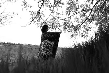 Black and white photograph of a girl holding an abstract-patterned carpet in wide open fields,...