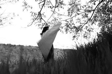 Black and white photograph of a girl holding an abstract-patterned carpet in wide open fields,...