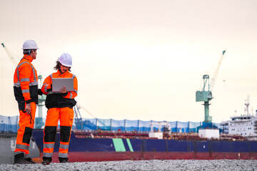 Fototapeta premium Two port engineers in high-visibility orange uniforms and hard hats communicate while inspecting dockside logistics using a laptop and walkie-talkie, symbolizing teamwork and safety.