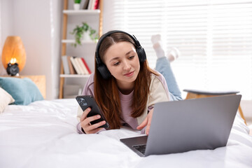 Teenage girl in headphones with smartphone and laptop on bed at home