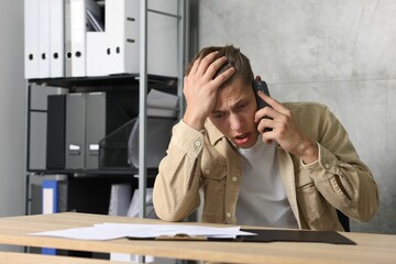 Young man with papers complaining to someone via phone at wooden table indoors