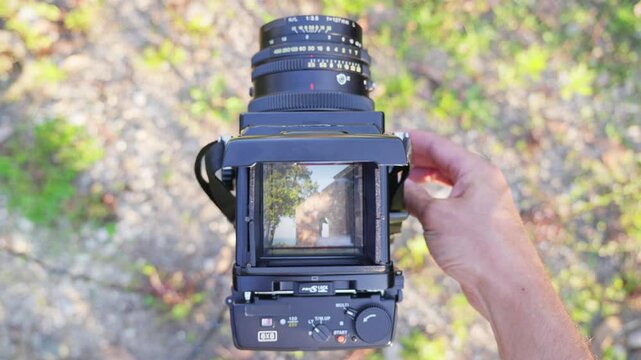 A man looks through the viewfinder of a vintage medium format camera. In the viewfinder, he sees the antique building on a sunny day