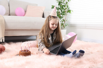 Birthday celebration. Little girl with cake having video call via laptop at home