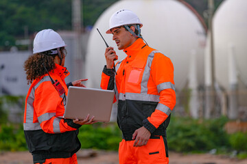 Two engineers in high-visibility safety gear and hard hats discuss project plans on a laptop in front of large spherical gas storage tanks at an industrial coastal facility.
