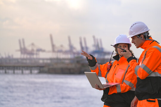 Two engineers wearing safety uniforms and hard hats communicate using a radio while reviewing data on a laptop by the sea during sunset. Maritime industrial setting.
