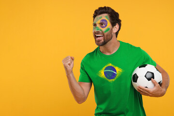 Excited fan with flags of Brazil on his face and t-shirt holding soccer ball against orange...