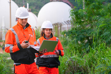 Two engineers in high-visibility safety gear and hard hats discuss project plans on a laptop in front of large spherical gas storage tanks at an industrial coastal facility.