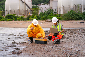 Two environmental engineers in safety gear work at a shoreline near gas storage tanks. One collects water samples, while the other records data on a laptop during site inspection.