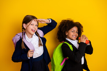 Two cheerful schoolgirls in uniforms with backpacks posing against a vibrant yellow background