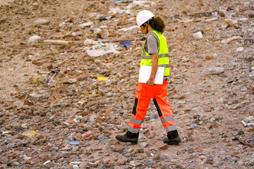 Female environmental engineer surveys rocky shoreline littered with waste, holding a laptop to...