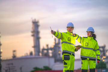 Two engineers in high-visibility suits and helmets review data on a tablet while standing in front of a refinery at sunrise, indicating teamwork and safety in a heavy industry zone.