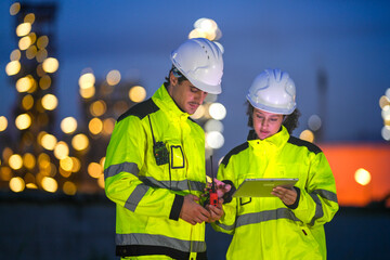 Two engineers in reflective safety gear review plans on a tablet during a night shift at an...