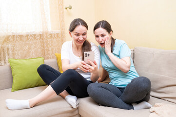Two female friends laughing on the couch while looking at a smartphone, discussing something funny,...