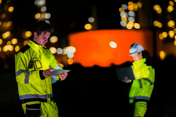 Two engineers collaborate on a night shift using a laptop and tablet at a plant A concept for the...
