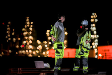 Two industrial workers take a break during the night shift at a construction site, sitting on steel...